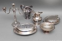 Photo of vintage silver plated serving pieces including oval chafing dishes, pitchers, ornate candelabra, and pierced bowl arranged on a gray surface.