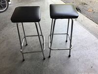 Two matching bar stools shown from front angle, black cushioned square seats, chrome legs with footrest bars, placed on concrete floor.