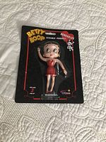 Front view of the Betty Boop bendable doll sealed in original black packaging with red and yellow print. Doll is in a red dress and poseable with bendable limbs.