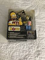 Front view of unopened Homer Simpson figure holding popcorn with packaging showing 'The Simpsons Movie' branding and sound feature indicated