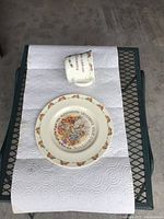 Photo showing both the cup and plate laid on paper towel on a metal mesh surface. The cup is tipped on its side displaying the Bunnykins Celebrate Your Christening colorful design on the body. The plate shows a central circular illustration with Bunnykins bunnies and celebratory flowers and text around the rim.