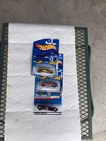 Three unopened Hot Wheels cars laid out on paper towel on a mesh table showing front packaging and car details.