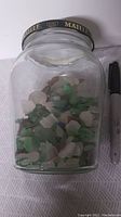 Front side view of a large clear glass jar labeled 'Maille 1747' black and gold lid filled about one-third with assorted green, white, and purple sea glass pieces. Sized next to a black marker for scale.