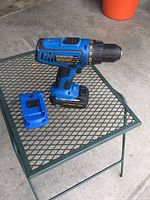 Blue and black Mastercraft cordless drill standing upright on a mesh table with its charger beside it.