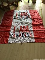 Two folded Canadian pole flags laid out on floor, showing 'I AM CANADIAN' text with maple leaf graphic, red side panels and central white panel