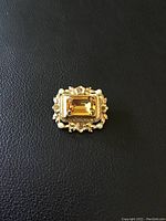 Front view of the vintage brooch displaying a large rectangular topaz centerpiece, framed by metal floral elements and small pearls on a black background.
