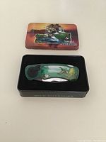 Closed folding pocket knife with green scenic wildlife design handle inside a black fitted tray within an illustrated tin box lid showing nature imagery and 'Wild Outdoors' text.
