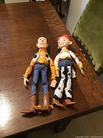 Woody and Jessie dolls lying on a wooden table, full body view showing clothes and boots, missing hats