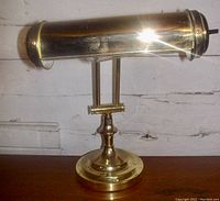 Front view of polished brass adjustable banker’s desk lamp with cylindrical shade and circular weighted base on wooden surface.