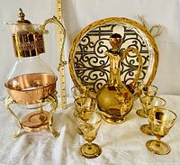 Photo showing entire lot with vintage glass coffee server in copper and metal stand on the left; gold-trimmed amber glass decanter and six matching glasses arranged in white wicker tray with black metal insert on the right. Ruler behind for scale.
