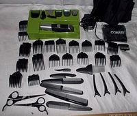 Top-down view showing the hair clipper in its green holder base, 14 black clipper guards arranged around, power cord, black carrying pouch, 4 hair clips, 6 combs, 2 scissors, and 2 mini brushes on a white textiles background.