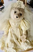 Close view of large teddy bear in ivory embroidered wedding gown, lace veil and tiara, original tags attached, showing front details.