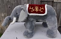 Large gray plush elephant with decorative red and gold saddle and white tusks, standing on white table outdoors.