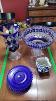 Top view of cobalt blue glassware including footed compote bowl, footed vase, blue plate, small vase, and candlesticks on wooden table.