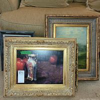 Three framed paintings arranged on carpet in front of couch showing still life with apples and vase, fox hunt scene, and landscape.