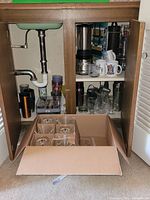 Open cabinet showing blender, mugs, shaker, stirrers, and glassware with a cardboard box containing six stemware glasses on floor in front.