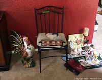 Photo of chair with cushion, large ceramic rooster figurine, placemats, napkin holder, candle holder, and slate art arranged on floor against red wall.