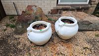 Photo showing the two matching concrete flower pots placed outdoors on gravel near large rocks, highlighting the overall shape, condition, and decorative elements.