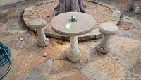 Concrete round table with twisted pedestal base and two matching textured concrete stools shown outdoors on a stone floor near a tree