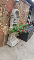 Full view of faux concrete garden fountain installed next to stone wall and black container, showing base and flower planter with vinca flowers.