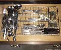 Silver and stainless steel flatware sorted in a wire tray showing spoons, forks and knives with varying handle designs.