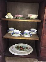 Porcelain teacups with saucers and three decorative plates featuring floral and fruit designs, arranged on wooden shelves.