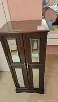 Front view of tall wooden jewelry cabinet with two mirrored paneled doors and metal handles.