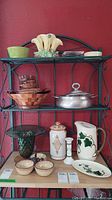 Display of various vintage serving pieces on a green metal shelf against red wall, showing a range of pottery, wood, glass, and pewter items.