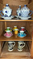 Wide photo showing all items in the set on wooden shelving: three Bombay lidded cups/saucers, six colorful tea cups/saucers, and two ceramic stein mugs