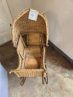 Front view of the antique wicker doll carriage showing the seating area, curved wicker sides and front, and wooden slats at the base.