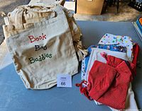 Photo showing stack of beige canvas drawstring tote bags with 'Book Bag Buddies' printed text and assorted colorful drawstring bags with different patterns including polka dots and floral.
