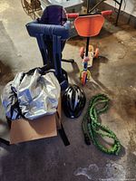 Photo shows a folded silver car cover on the left, a black bicycle helmet placed in front, a navy blue toddler bicycle seat standing upright behind, and a three-wheeled children's scooter with red hand grips and orange front shield on the right.