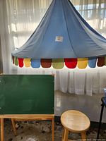 Blue fabric hanging canopy with multicolored scalloped edge shown with round wooden stool and green chalkboard easel in background.