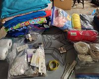 Various wool and fleece fabrics stacked, assorted yarn bundles wrapped in plastic, an assortment of sewing notions and crafting supplies spread out including buttons, trims, tape.