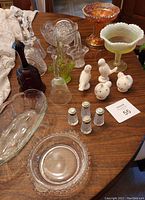 Photo showing a collection of crystal and glass dishes, various colors and styles, white ceramic bird figurines, and four small glass salt and pepper shakers on wooden table.