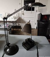 Two black desk lamps on a table. One lamp has an adjustable arm and round base; the other has a compartmentalized rectangular base and a conical shade.
