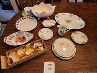 Collection of assorted dishes and glassware displayed on a wooden table including plates, bowls, pitchers, and a box of glassware with gold accents.