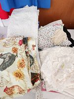 Image showing folded white lace tablecloths, a fabric with rooster and sunflower print, floral fabric, and vintage knit gloves on top