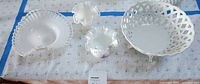 Top-down photo showing four milk white glass dishes arranged on fabric. Items include footed lattice basket, two ruffled edge bowls, and divided dish. The dishes have glossy opaque white finish with no chips visible.