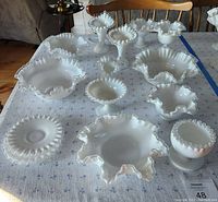 Photo of 11 vintage milk white Fenton glass pieces on a cloth-covered table, showing various sizes of bowls, vases, and a covered jar all with ruffled or crimped edges.