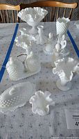Image showing a collection of 9 vintage Fenton milk white glass items on a table, featuring various bowl sizes, a tall vase, candle holders, a covered butter dish, and decorative hobnail textured pieces with ruffled edges.