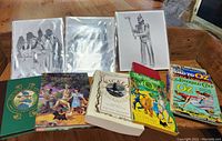 Shows six Wizard of Oz books laid side by side on a wooden surface with three framed black and white sketches leaning behind them.