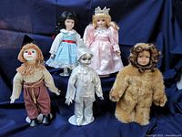 Photo showing five Seymour Mann Wizard of Oz dolls arranged against a dark blue fabric background: the Scarecrow, Dorothy, Good Witch, Tinman, and Lion.