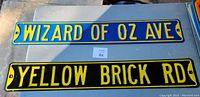 Photo showing two blue metal street signs with yellow lettering reading 'WIZARD OF OZ AVE' and 'YELLOW BRICK RD.' The signs are laid on a flat surface with good visibility.