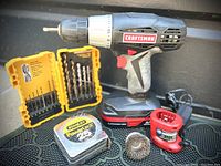 Photo showing Craftsman cordless drill with attached battery, yellow drill bit case, Stanley tape measure, wire brush, red battery charger.