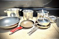 Wide shot of eight cookware items: three stainless steel saucepans with lids, large stock pots, non-stick frying pan with red handle, non-stick wok