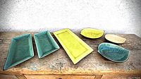 Six glazed ceramic serving dishes arranged on wooden surface: 3 rectangular trays (green, yellow, blue-green) and 3 leaf-shaped bowls (green, yellow, beige).