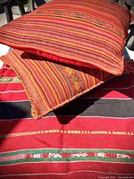 Close-up of two hand-woven pillows with red, orange, and yellow tones lying on the tapestry throw with red, black, green and white stripes and embroidery.