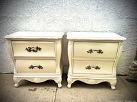 Front view of the pair of reproduction French Louis XVI style nightstands showing their off-white finish and 2 drawers with decorative metal pulls.