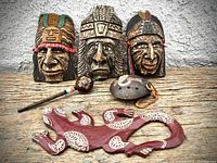 Front view of three Native American chieftain carved wooden masks, a rattle, a flute, and a maroon gecko wood carving on a rustic wooden surface.